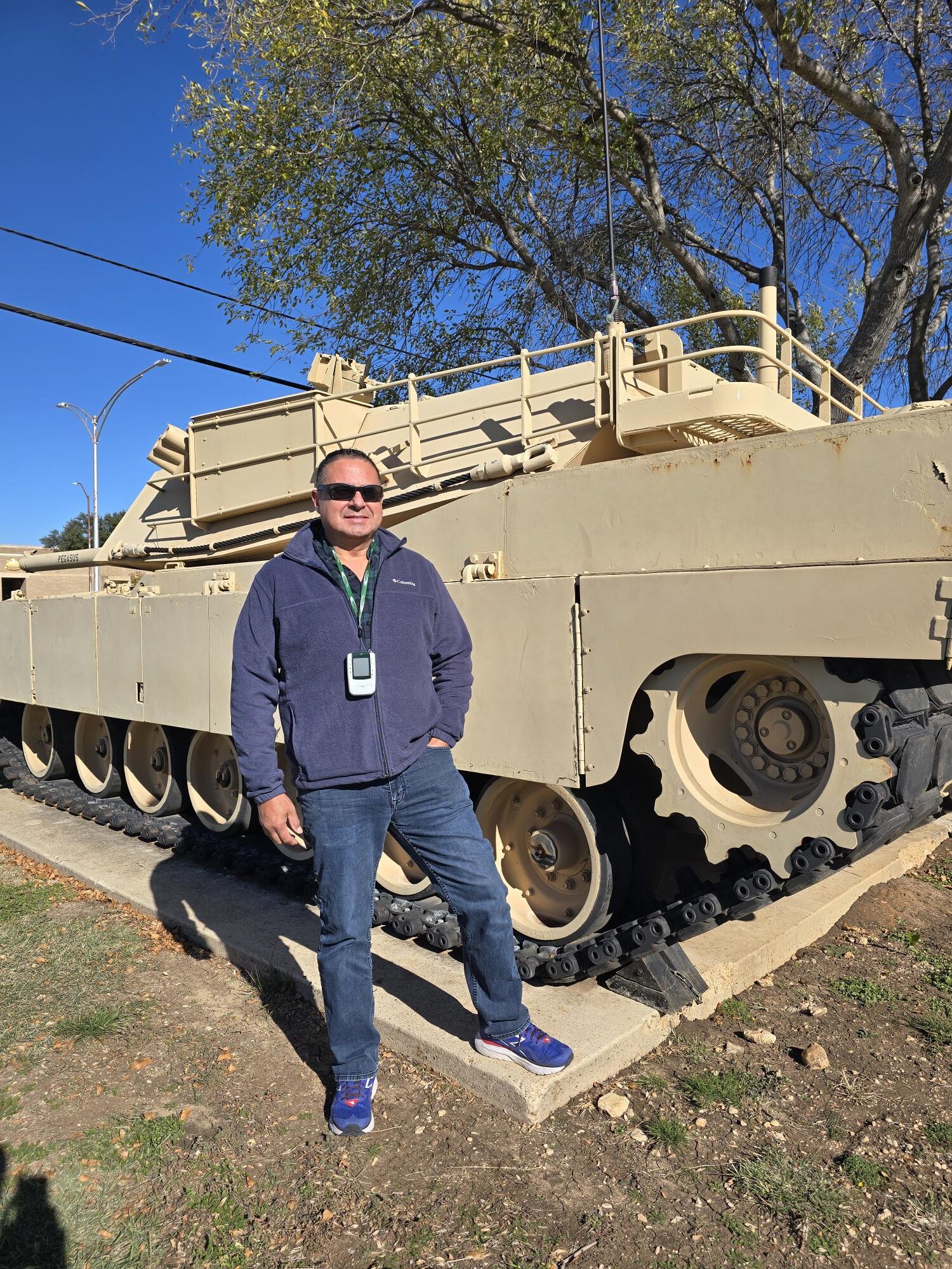 Lieutenant Colonel in sunglasses and a blue jacket stands in front of a large tan military tank outdoors on a sunny day.
